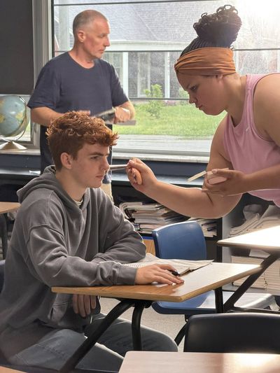 A teenage boy sits at a classroom desk while a makeup artist leans in to apply makeup to his face. An older man stands in the background near a window, reviewing notes. Natural light fills the classroom.