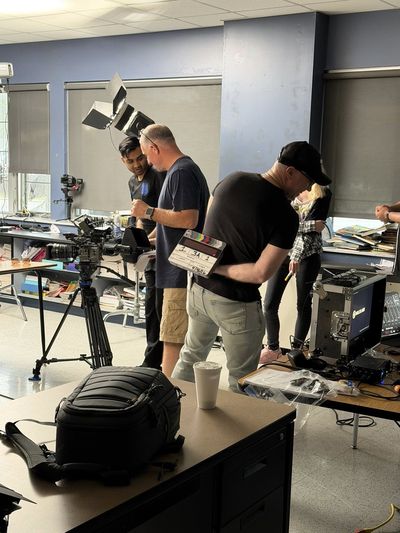 Three crew members stand in a classroom surrounded by production equipment including a camera on a tripod, a large light with barn doors, and a sound mixing board. One crew member holds a film slate.