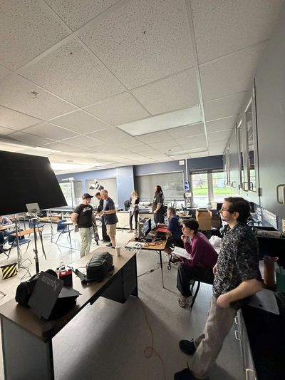 A large classroom has been transformed into a film production space. Multiple crew members work at tables covered in production equipment, monitors, and gear. Large lights and camera rigs line the walls.