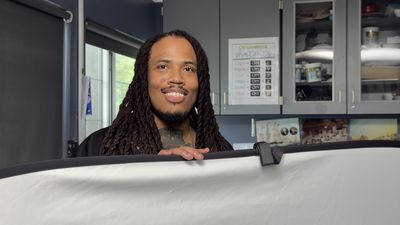 A man with long dreadlocks and a warm smile leans over a large white reflector panel on set. Blue-gray cabinetry and a Chromebook shortcuts poster are visible in the background.