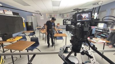 A professional cinema camera on a slider in the foreground with a live monitor attached. Behind it a crew member walks between classroom desks adjusting the setup while two teenage boys sit waiting in position.