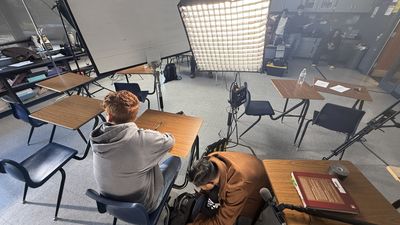 A large square softbox light dominates the centre of a classroom while a teenage boy sits at a desk in the foreground with his back to the camera. Crew work in the background near production equipment.