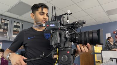 A young man holds and adjusts a large Sony cinema camera rigged with a wireless transmitter, antenna array, and cinema lens. A classroom with blue-gray cabinetry is visible behind him.