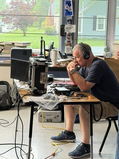A man sits at a desk covered in production equipment, monitors, and cables, wearing headphones and leaning forward with his hands clasped in concentration. A window looks out on a green suburban street.