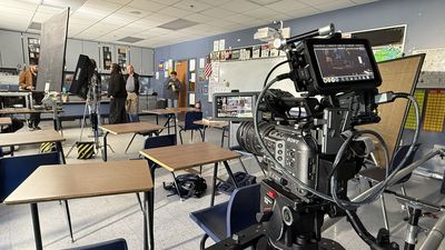 A Sony cinema camera with dual monitors in the foreground. Behind it, several crew members and actors stand in conversation in a classroom between takes.