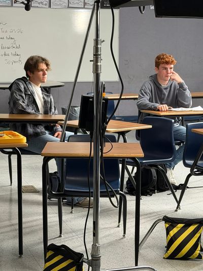Two teenage boys sit at separate desks in a classroom, waiting between takes. A light stand and sandbags are visible in the foreground, a whiteboard with a class agenda on the wall behind them.