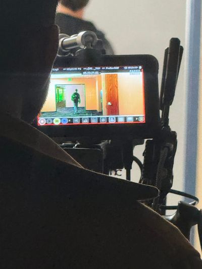 A camera operator in silhouette in the foreground, with a small on-camera monitor showing a figure walking down a hallway toward a door.