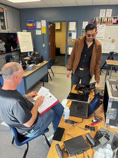 A man sits in a classroom chair reviewing a script binder while another stands nearby at a desk covered in production equipment, a laptop, wireless transmitters, and cables.