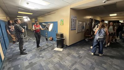 Three crew members adjust lighting equipment in a school hallway lined with blue lockers. A portable light on a stand is being positioned centre frame while teenage extras wait along the right.