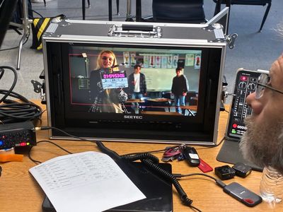 A large production monitor mounted in a travel case displays a live camera feed of a woman holding a film slate in front of two teenage boys. Cables, a sound mixer, and a script binder sit on the desk in front of it.