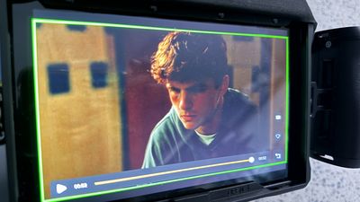 A mobile director's monitor displays a close-up playback of a teenage boy with his head bowed, lit by warm amber light against a blurred background of school lockers.