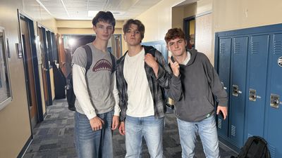Three teenage boys stand together in a school hallway lined with blue lockers, smiling and relaxed between takes. A production light glows in the background.