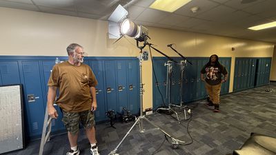 A man stands in a school hallway lined with blue lockers next to a tall production light on a stand and a boom microphone arm. A second man watches from further down the hallway.