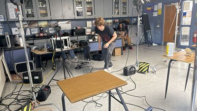 A young crew member caught mid-motion in a classroom surrounded by production equipment, light stands, cables, and battery packs, with two others working at a production desk behind.