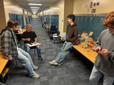 Four people sit and stand in a long school hallway lined with blue lockers. A man seated in the centre holds an open script binder while three younger men gather around him in conversation.