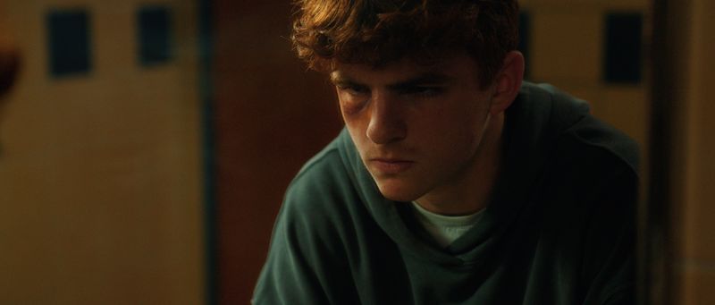 Close on a teenage boy in a green shirt, head lowered, lit from the side.