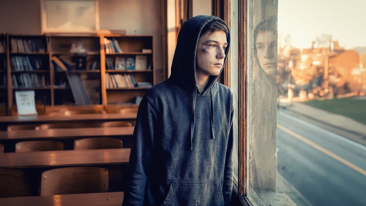 A teenager in a hoodie looks out a school window at dusk.