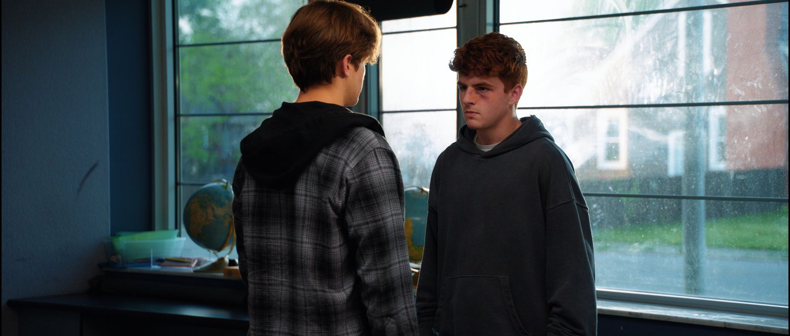 Two teenage boys stand face to face in front of a bank of classroom windows.