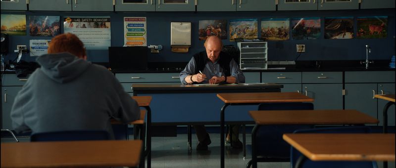 A teacher sits marking papers at the front of an empty classroom while a student sits at the back.