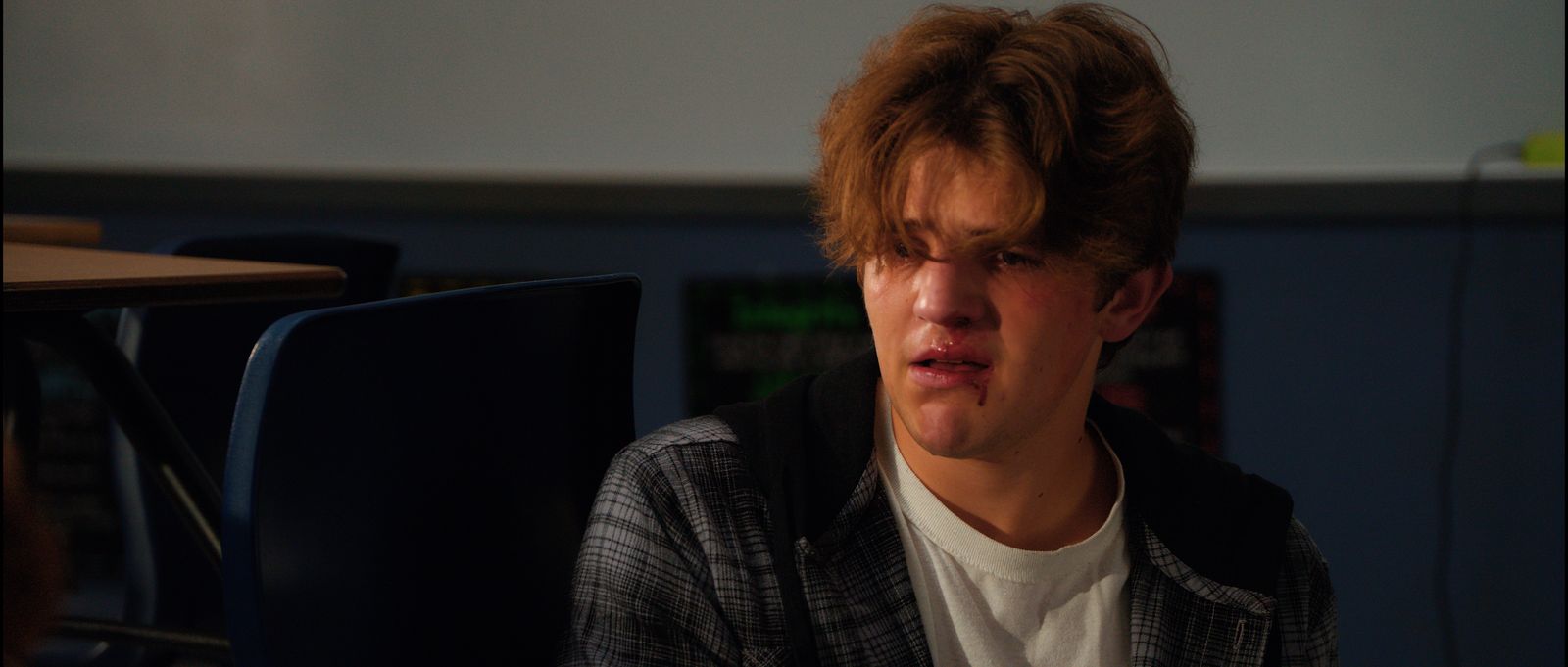 A teenage boy sits at a desk, eyes red, staring ahead.