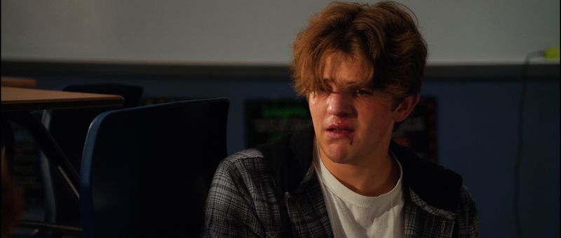 A teenage boy sits at a desk, eyes red, staring ahead.
