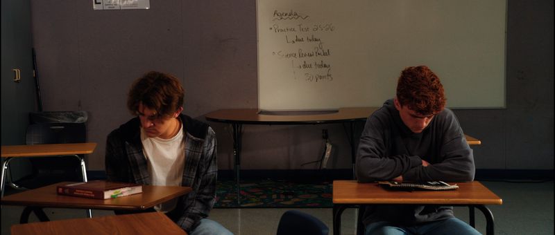 Two teenage boys sit at opposite desks in an empty classroom, both looking down.