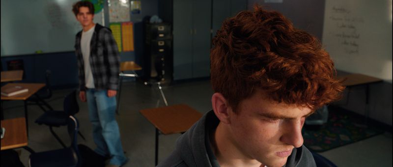 A teenage boy in the foreground looks down while another watches from across the classroom.